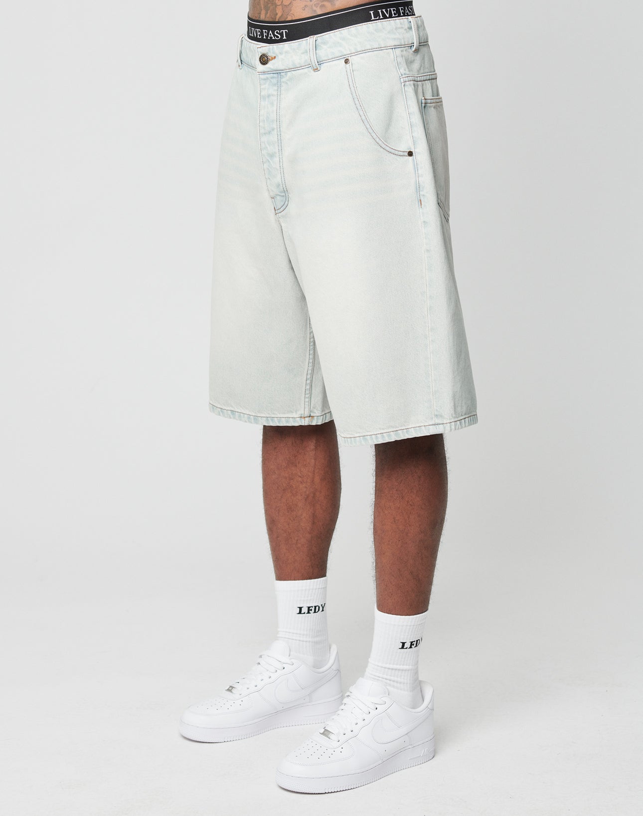 A person wears LIVE FAST DIE YOUNG LFDY Jorts—light-wash, baggy denim shorts with a back pocket—paired with white LFDY socks and white Nike sneakers. Only the lower half of their body is shown against a plain white background.