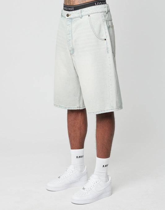 A person wears LIVE FAST DIE YOUNG LFDY Jorts—light-wash, baggy denim shorts with a back pocket—paired with white LFDY socks and white Nike sneakers. Only the lower half of their body is shown against a plain white background. | model