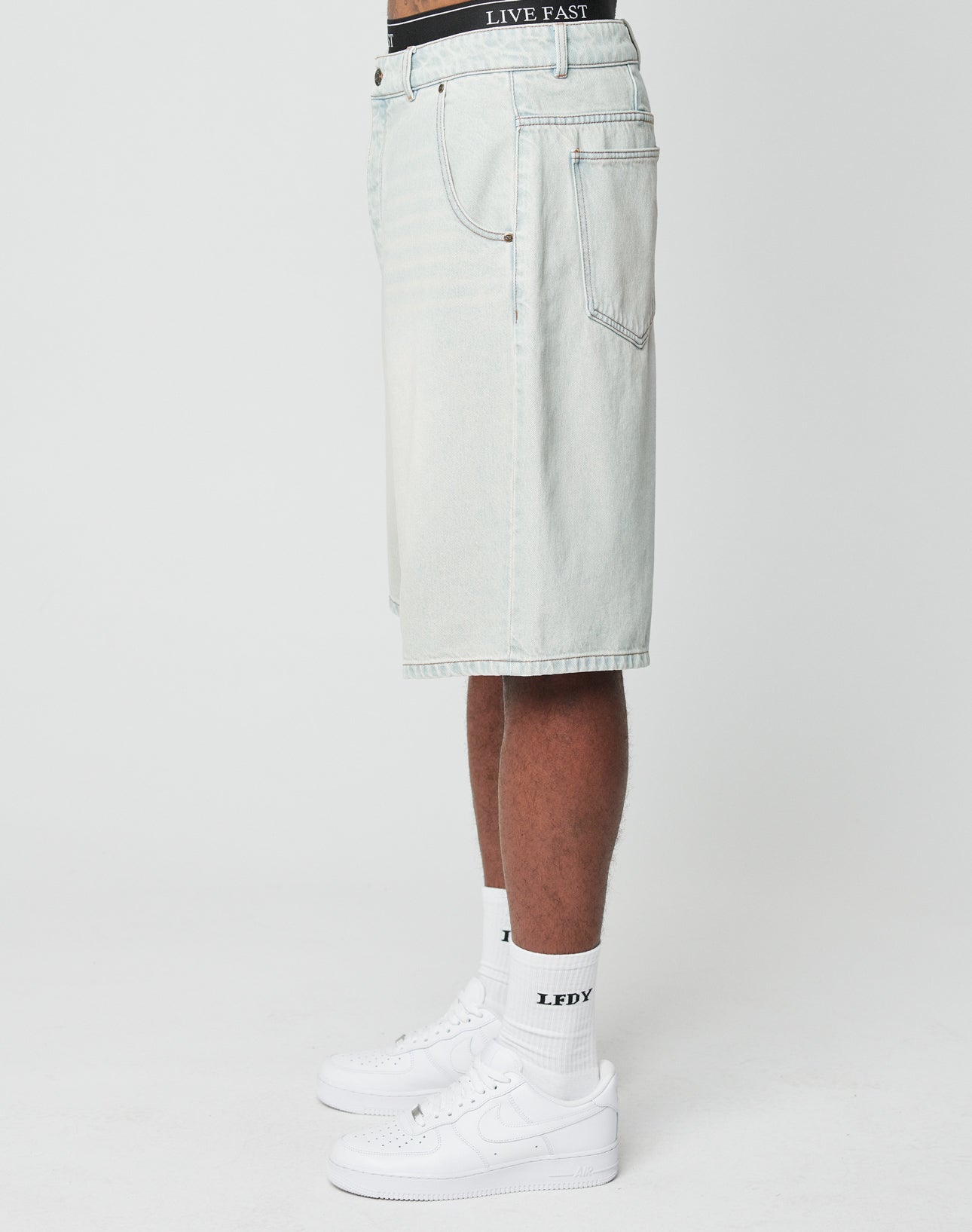 A person stands in profile against a plain white background wearing LIVE FAST DIE YOUNG LFDY Jorts with back pockets, white Nike sneakers, white socks with LFDY text, and a LIVE FAST waistband visible above the shorts.