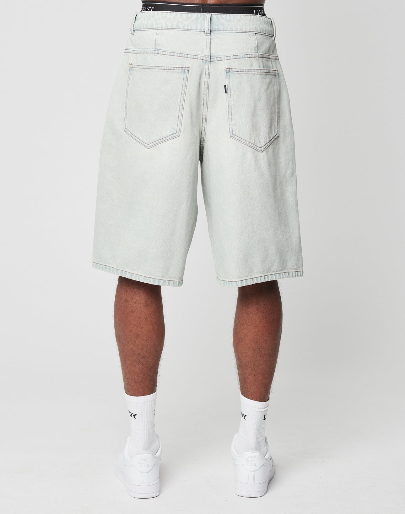 Someone is shown from the back wearing LIVE FAST DIE YOUNG LFDY Jorts—light-wash, knee-length denim shorts with a Gesäßtasche—paired with white socks and sneakers, standing against a plain white background.