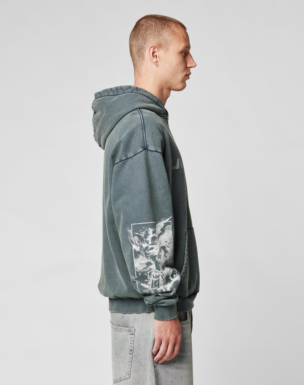 A young man in profile wears the LFDY Window Hooded by LIVE FAST DIE YOUNG—a faded, dark green hoodie with a reflective front print and white sleeve graphic—paired with light gray jeans against a plain, light background.