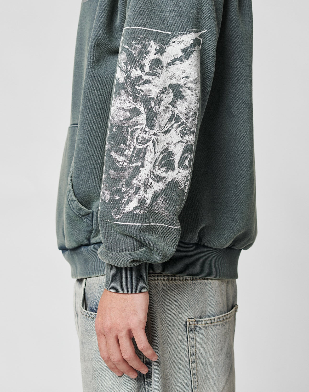 A person wears the LFDY Window Hooded sweatshirt by LIVE FAST DIE YOUNG, featuring an oversized green fit with a reflective front print and a white abstract graphic on the sleeve, paired with light blue jeans. Only the torso, part of one arm, and hand are visible.