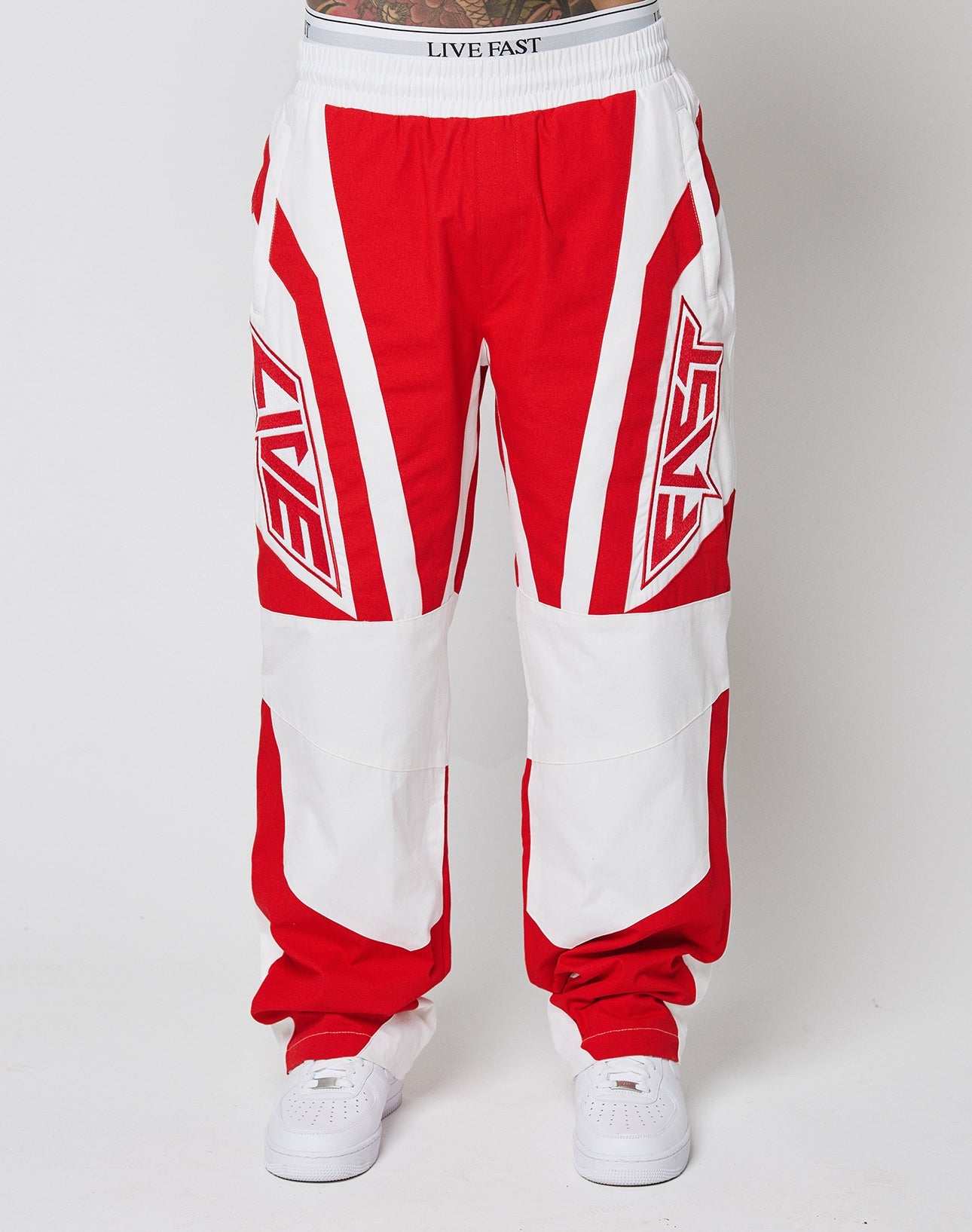 A person wears LFDY Cross Pants by LIVE FAST DIE YOUNG—red and white with bold geometric patterns, Stick Applikationen, and LIVE FAST on the front and waistband. Made from 100% Baumwolle, paired with white sneakers.