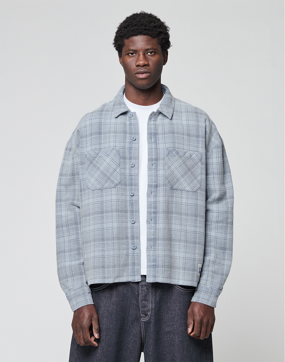 A man faces forward wearing a light gray plaid Vintage Flannel by LIVE FAST DIE YOUNG, styled in an oversize fit over a white t-shirt and dark jeans, set against a plain light background. | model