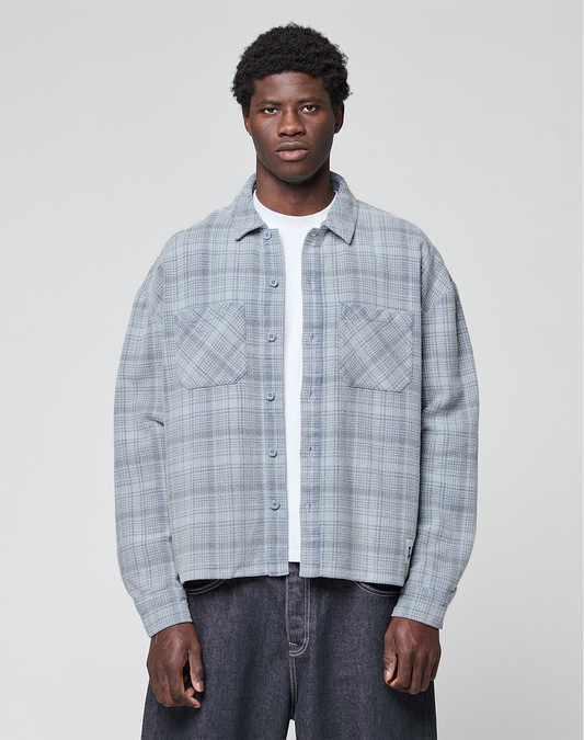 A man faces forward wearing a light gray plaid Vintage Flannel by LIVE FAST DIE YOUNG, styled in an oversize fit over a white t-shirt and dark jeans, set against a plain light background. | model