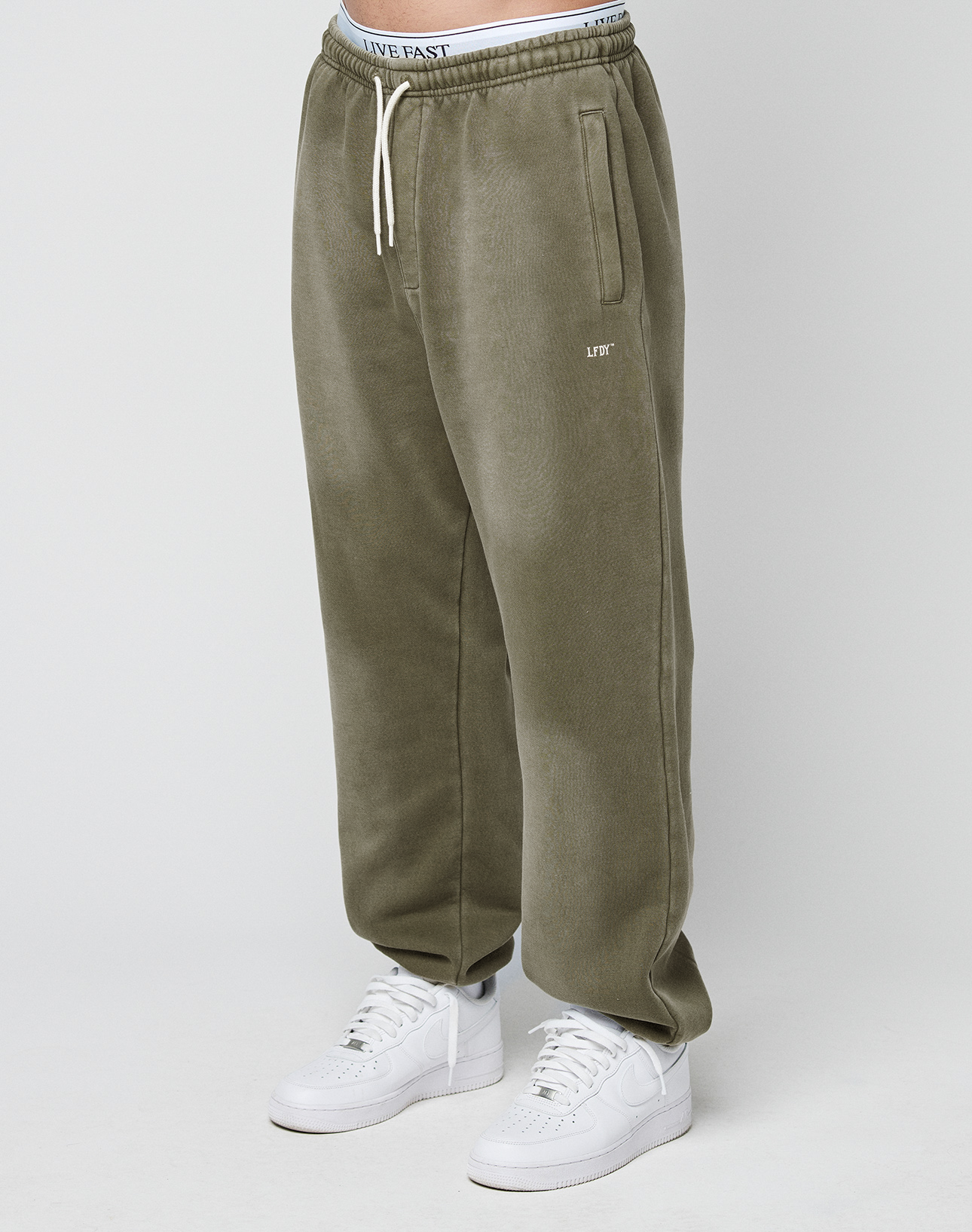 A person wears the Basic Loose Jogger by LIVE FAST DIE YOUNG in olive green with a drawstring waist, white LIVE text on the left thigh, and an elastic waistband with LIVE FAST text, styled with white sneakers on a plain white background. | model
