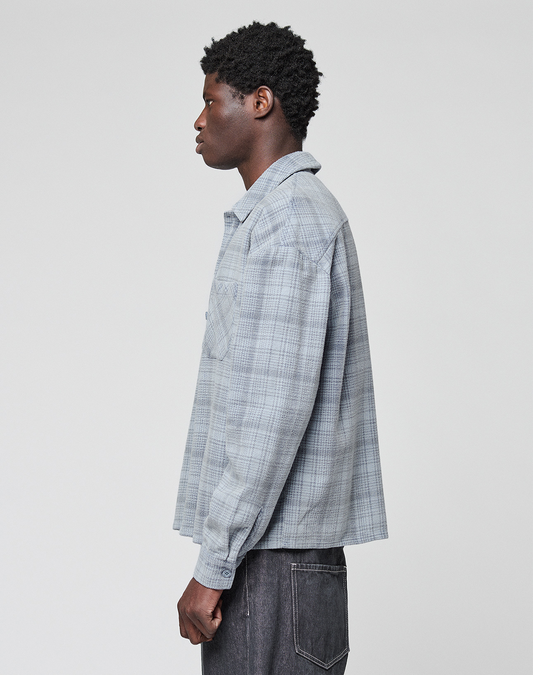 A person with short curly hair stands in profile wearing the LIVE FAST DIE YOUNG Vintage Flannel in light gray plaid and dark jeans against a plain light background.
