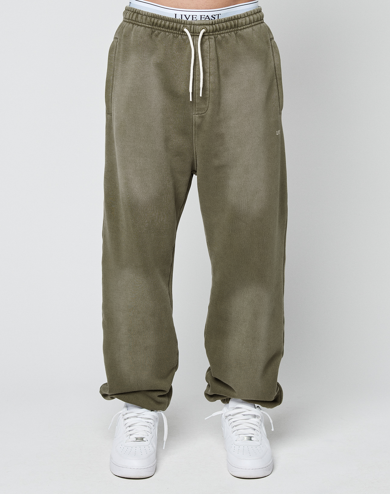 A person wears the LIVE FAST DIE YOUNG Basic Loose Jogger in olive green with white drawstrings, paired with white sneakers and visible LIVE FAST waistband. The joggers have a subtle faded look, shown against a plain, light background.