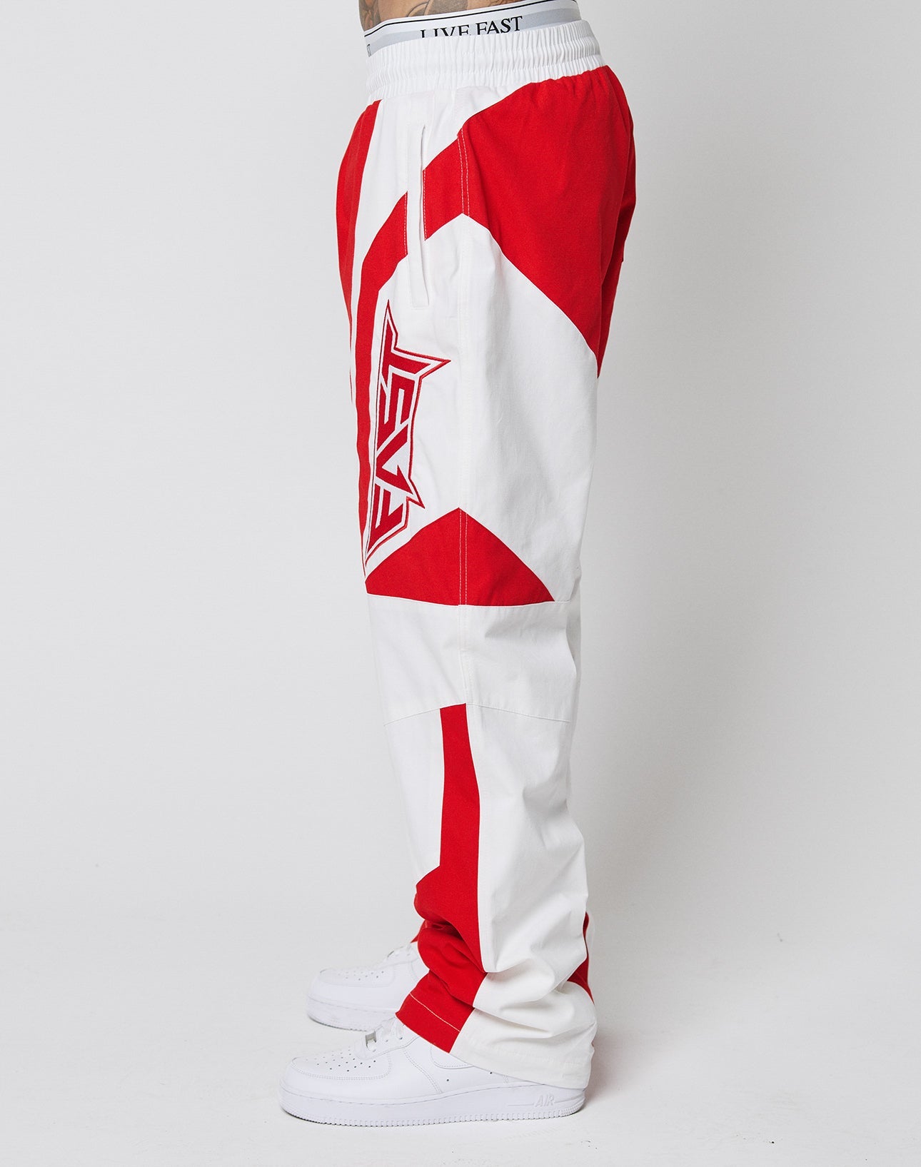 A person in oversize fit LFDY Cross Pants by LIVE FAST DIE YOUNG, featuring bold white and red graphics and LIVE FAST on the waistband, stands in side profile with white sneakers against a plain white background.