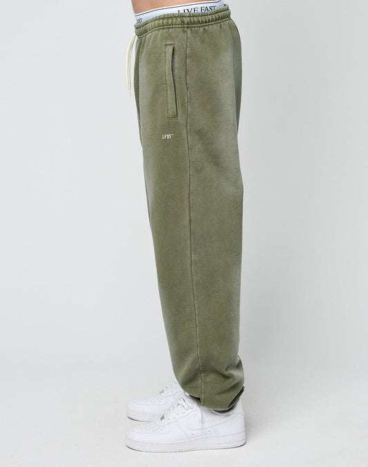 A person stands sideways against a plain light background wearing LIVE FAST DIE YOUNG Basic Loose Jogger sweatpants in olive green with zipper pockets, logo print detail, a white LIVE FAST waistband, and white sneakers.