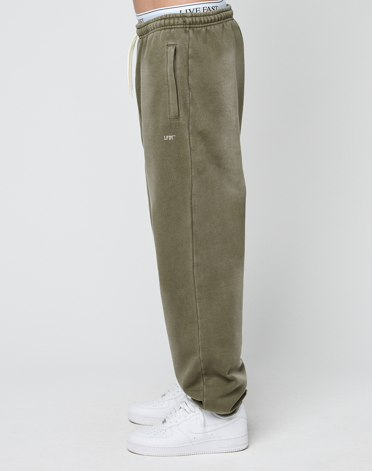 Model wears Basic Loose Jogger by LIVE FAST DIE YOUNG in olive green with handgefertigtem Fade-Effekt, LFYT text on the upper thigh, white drawstring, and white sneakers while standing sideways against a plain white background.