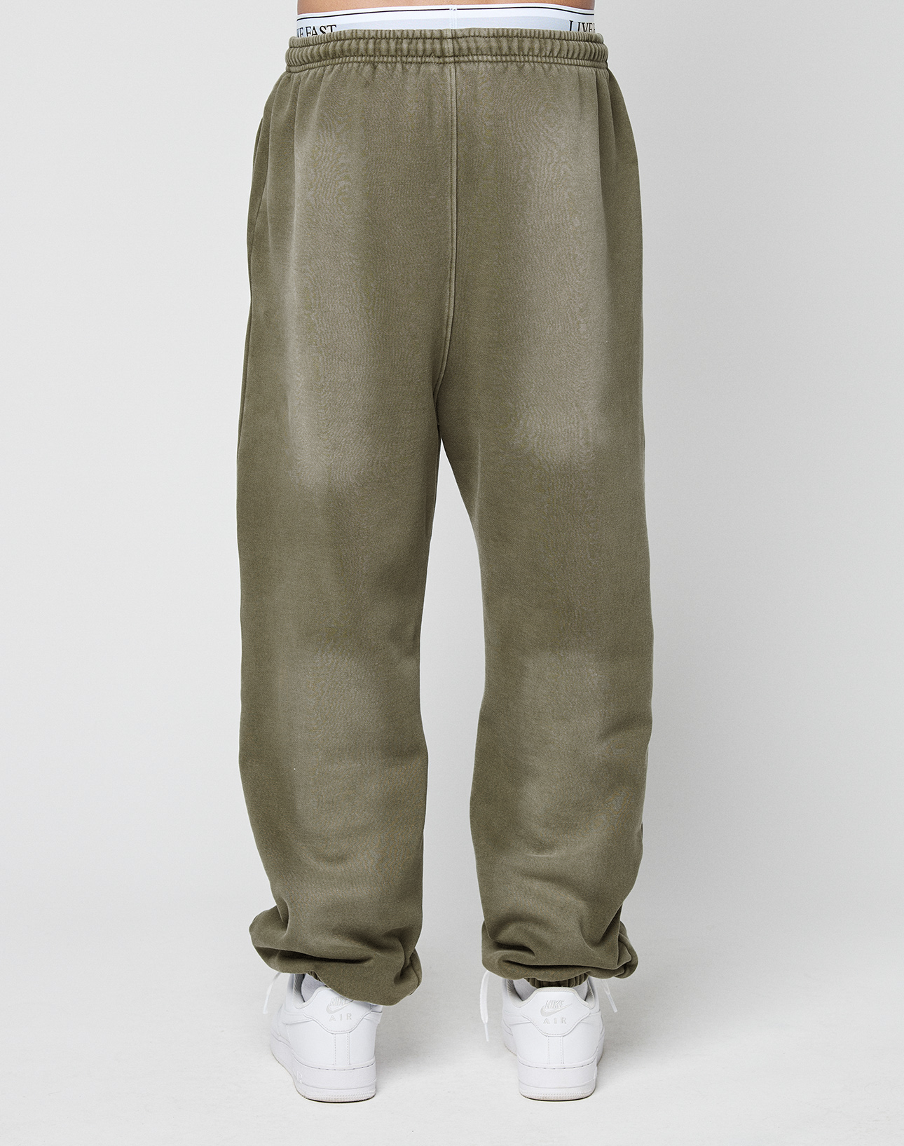 A person wears LIVE FAST DIE YOUNG’s Basic Loose Jogger in olive green with an elastic waistband and subtle faded look, white sneakers, and partially visible white underwear, standing against a plain white background.