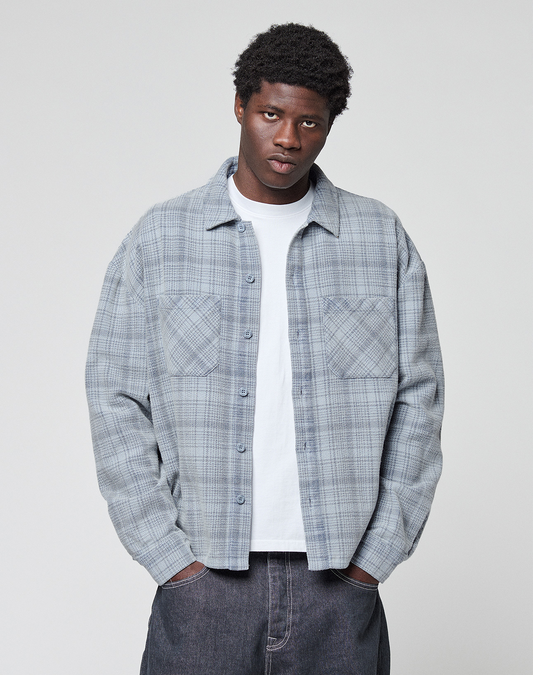 A person wearing an oversized Vintage Flannel by LIVE FAST DIE YOUNG in light gray plaid over a white t-shirt stands with hands in pockets against a plain light background, looking directly at the camera.