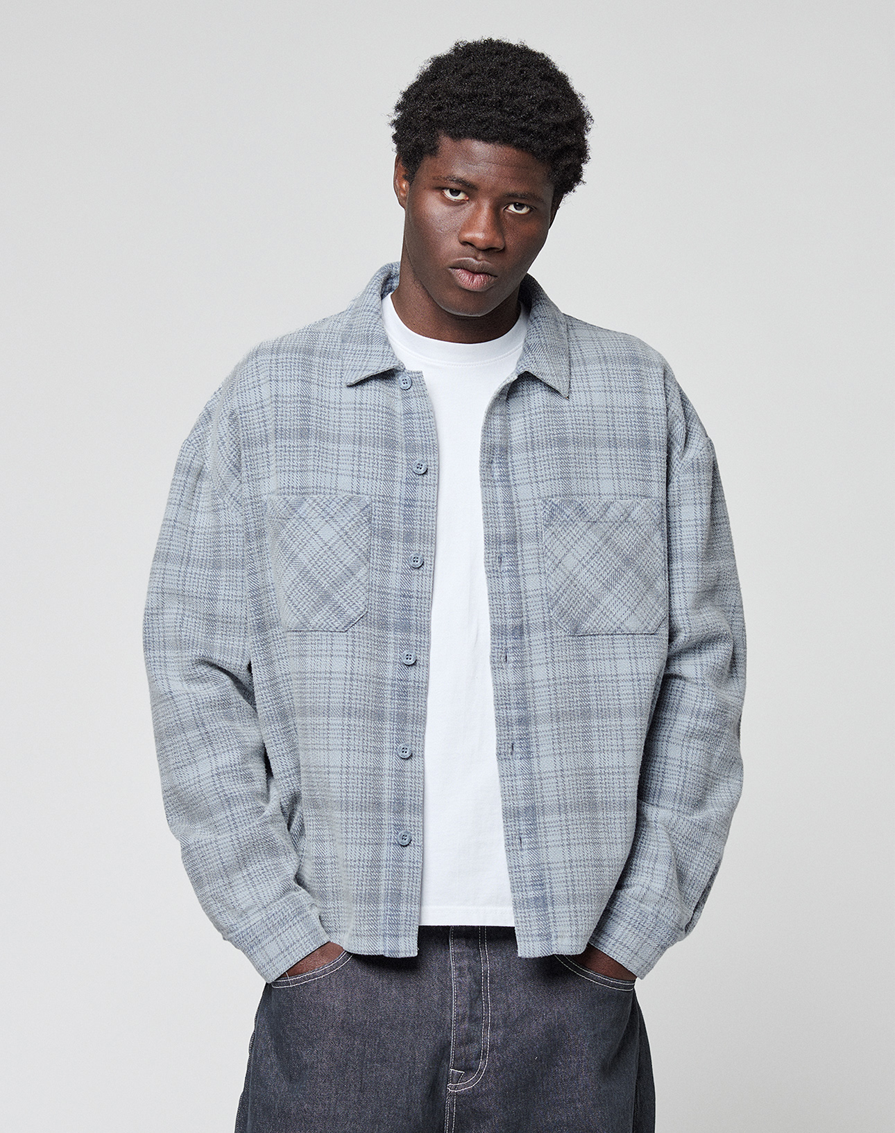 A person wearing an oversized Vintage Flannel by LIVE FAST DIE YOUNG in light gray plaid over a white t-shirt stands with hands in pockets against a plain light background, looking directly at the camera.