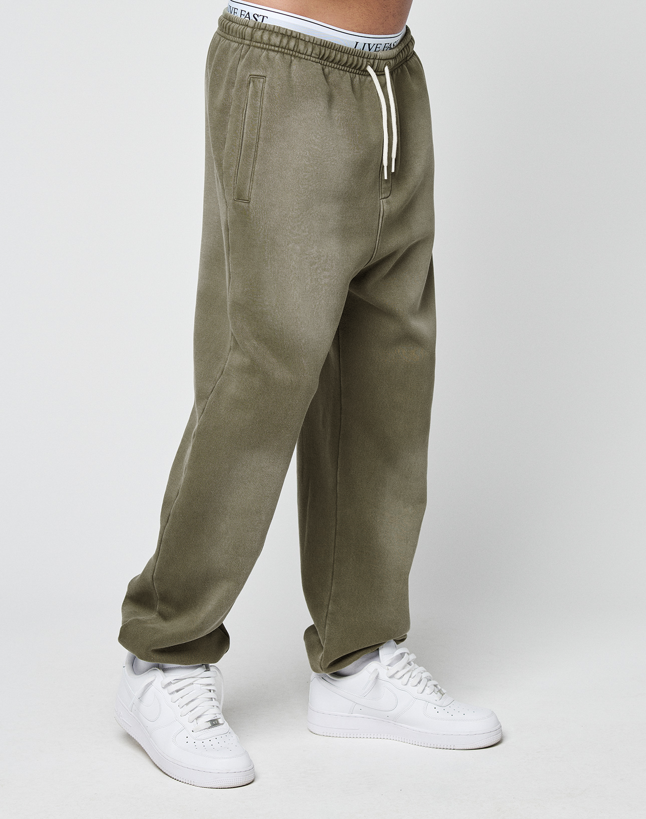 A person is shown from the waist down wearing LIVE FAST DIE YOUNG Basic Loose Jogger sweatpants in olive green with white drawstrings and white sneakers, standing against a plain white background.