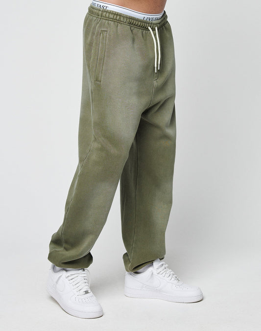 The lower half of a person is shown wearing LIVE FAST DIE YOUNG Basic Loose Jogger sweatpants in olive green with white drawstrings and white sneakers, standing against a plain light background.