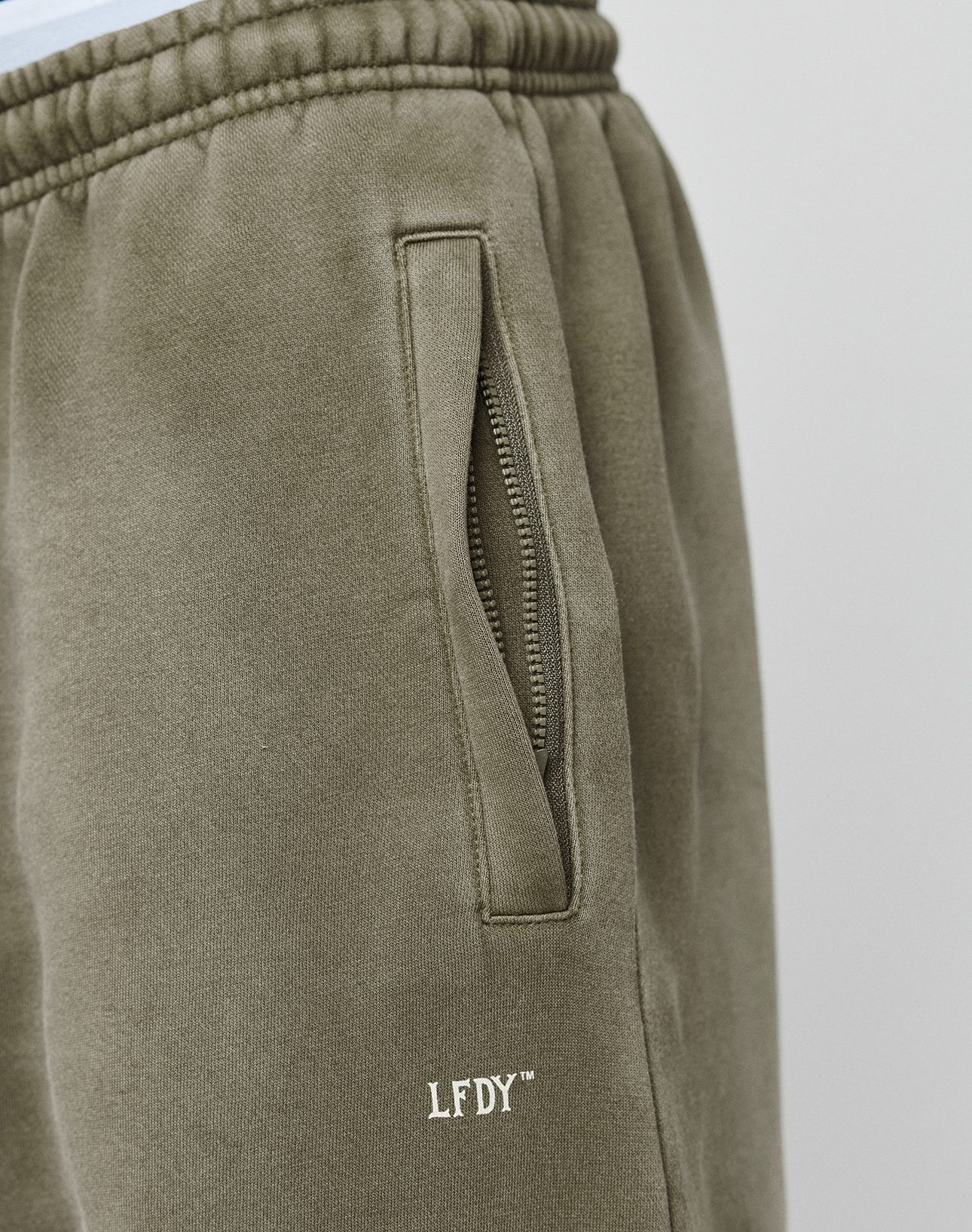 Close-up of the LIVE FAST DIE YOUNG Basic Loose Jogger in olive green, featuring a handmade fade effect, zippered side pocket, white LFDY logo near the pocket, loose fit, and elastic waistband. A white shirt is visible at the top.