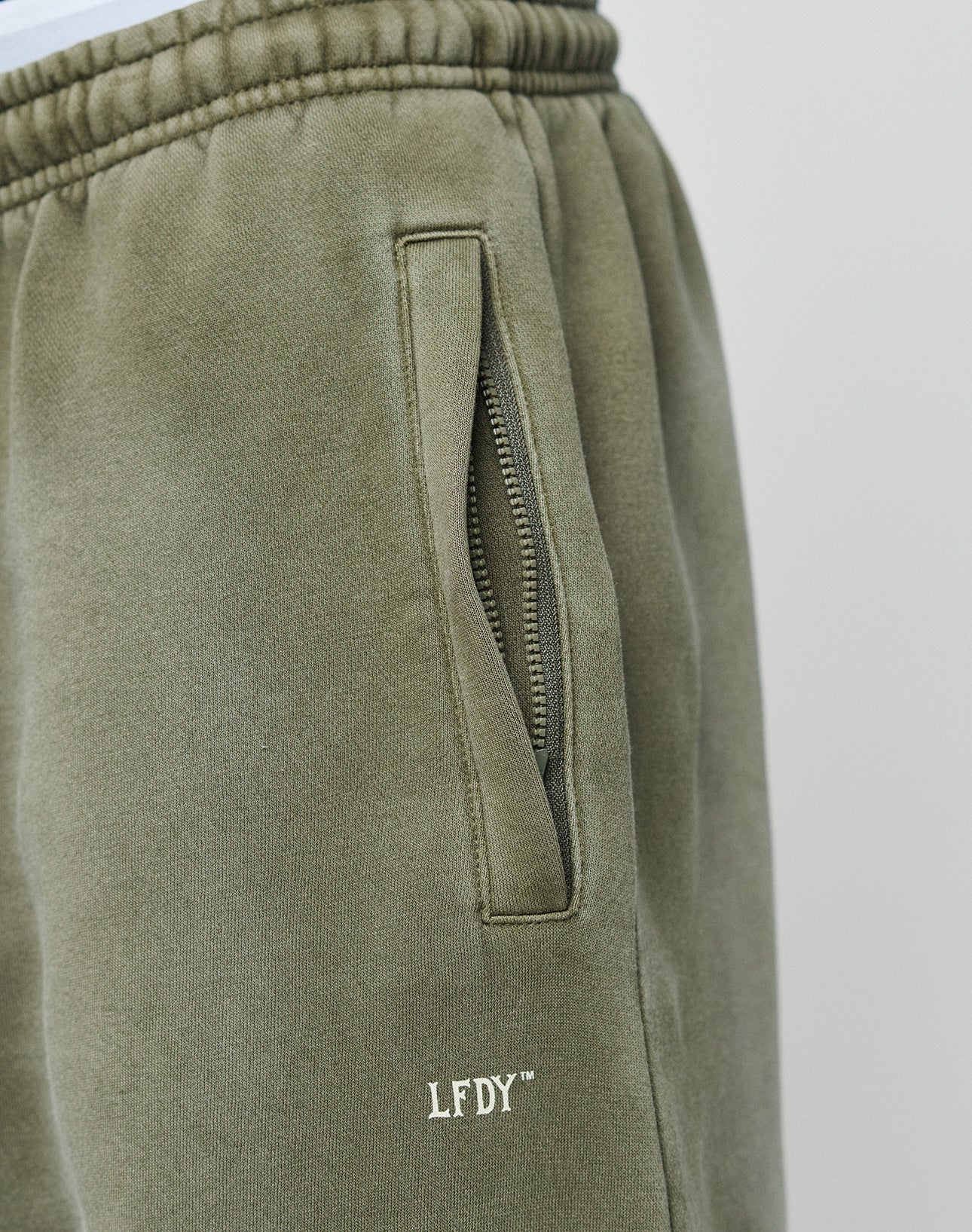 Close-up of LIVE FAST DIE YOUNGs Basic Loose Jogger in olive green, featuring a relaxed fit, zipper pocket, soft fabric with white LFDY™ embroidery near the pocket, visible stitching on the elastic waistband, and subtle logo print details.
