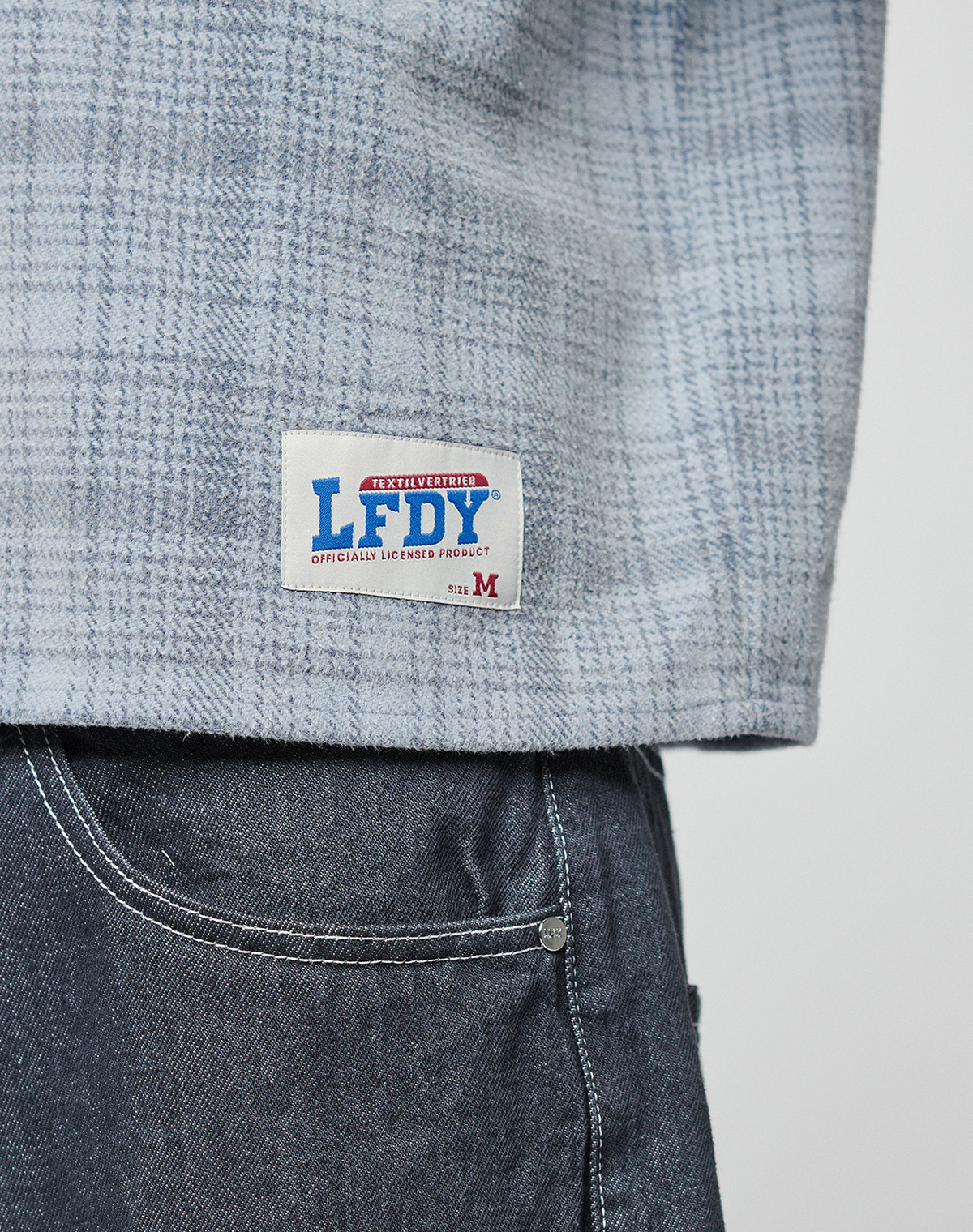 A close-up of a person wearing a LIVE FAST DIE YOUNG Vintage Flannel in light blue plaid, oversize fit, with the brand patch on the hem and medium-wash jeans. Label: OFFICIALLY LICENSED PRODUCT, size M.