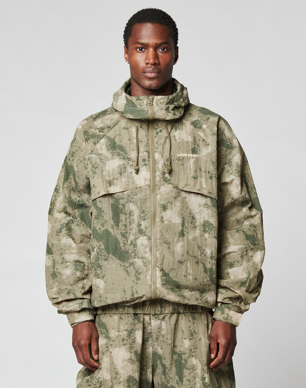 A man stands facing forward in the LIVE FAST DIE YOUNG Live Fast Nylon Jacket, featuring a green and beige camo pattern, oversize fit, front zipper, two large chest pockets, a hood with elastic drawcord, and matching pants. | model