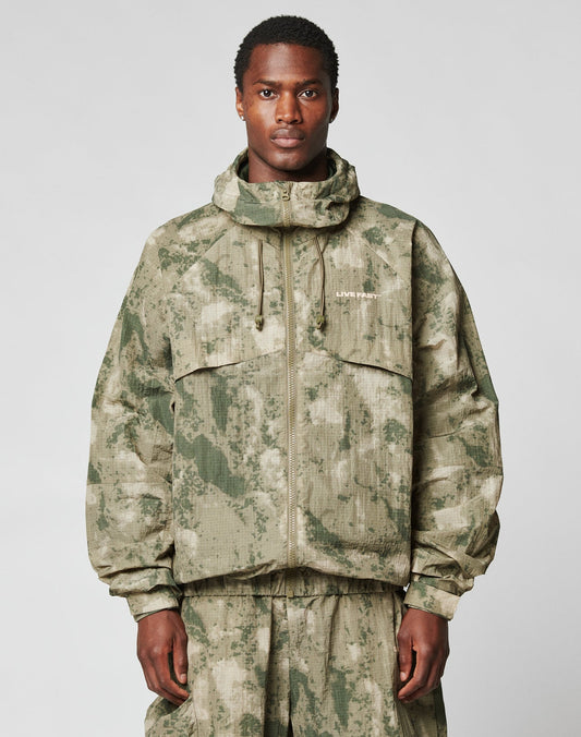 A man stands facing forward in the LIVE FAST DIE YOUNG Live Fast Nylon Jacket, featuring a green and beige camo pattern, oversize fit, front zipper, two large chest pockets, a hood with elastic drawcord, and matching pants. | model