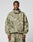 A man stands facing forward in the LIVE FAST DIE YOUNG Live Fast Nylon Jacket, featuring a green and beige camo pattern, oversize fit, front zipper, two large chest pockets, a hood with elastic drawcord, and matching pants.