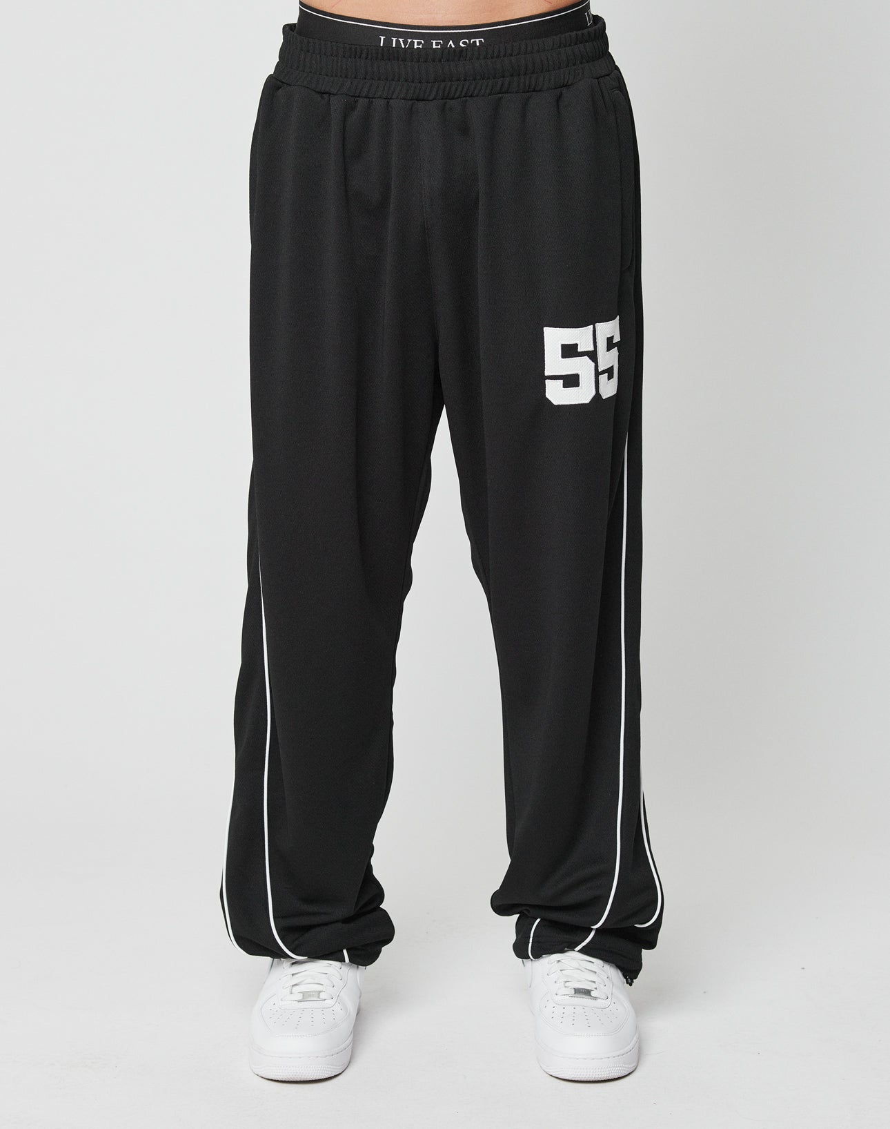 A person wears LFDY 55 Trackpants by LIVE FAST DIE YOUNG—loose black wide fit with a comfy elastic waist, white side piping, and “55” on the left thigh—paired with white sneakers, standing against a plain white background.
