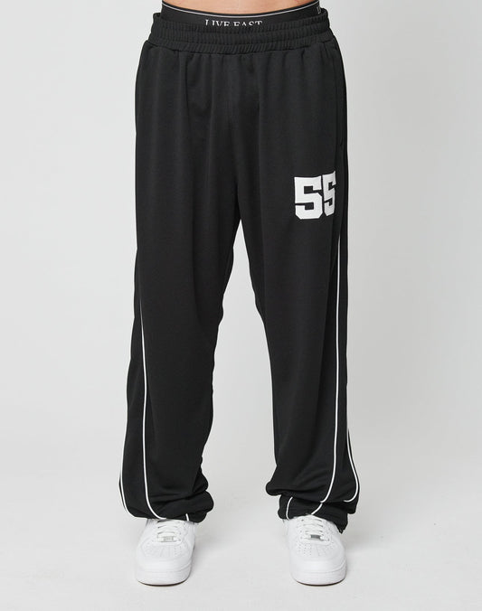 A person wears LFDY 55 Trackpants by LIVE FAST DIE YOUNG—loose black wide fit with a comfy elastic waist, white side piping, and “55” on the left thigh—paired with white sneakers, standing against a plain white background.