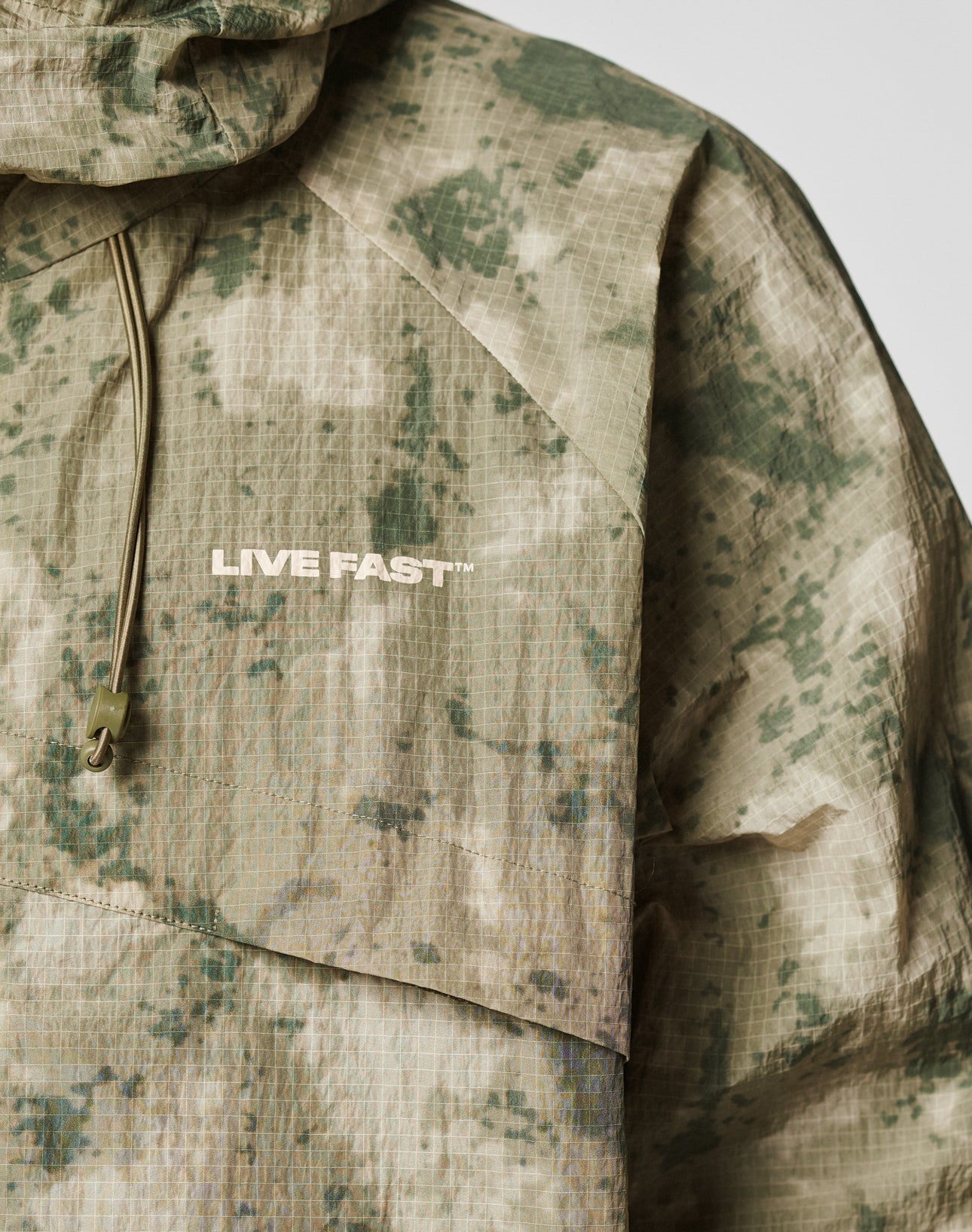A close-up of the LIVE FAST DIE YOUNG Live Fast Nylon Jacket, featuring green and beige digital camouflage, an oversize fit, elastic hood drawstring, and “LIVE FAST™” printed in white on the chest.