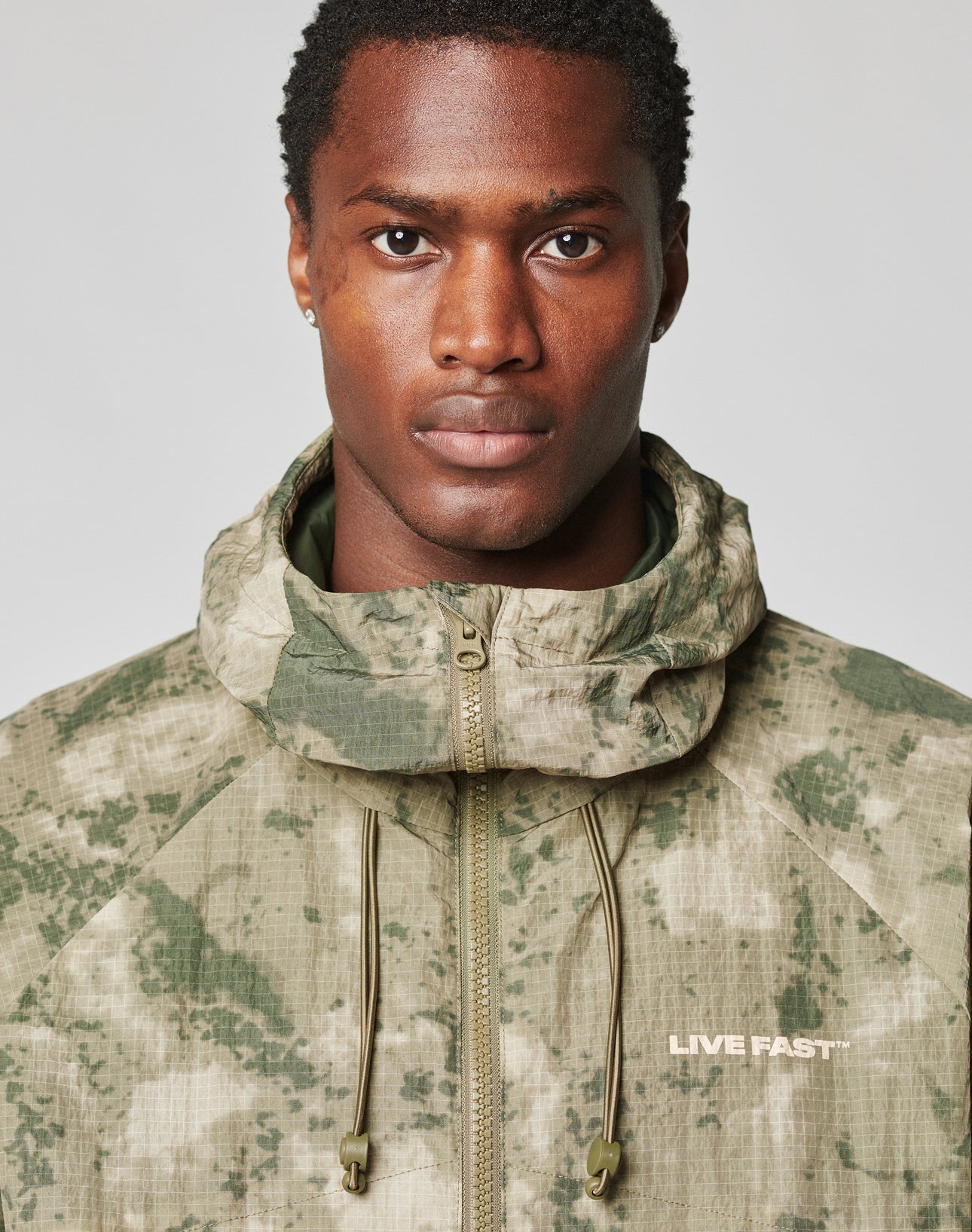 A man models the LIVE FAST DIE YOUNG Live Fast Nylon Jacket in green and beige camo, featuring a hood with elastic drawstring and LIVE FAST text on the chest, looking at the camera against a plain background.
