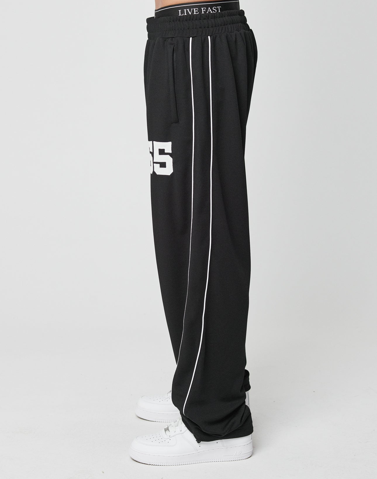 A person wears LIVE FAST DIE YOUNG LFDY 55 Trackpants featuring a wide fit, elastik-bund waist, white side piping, and 55 on the leg, paired with white sneakers, against a plain white background. Polyester fabric ensures comfort.