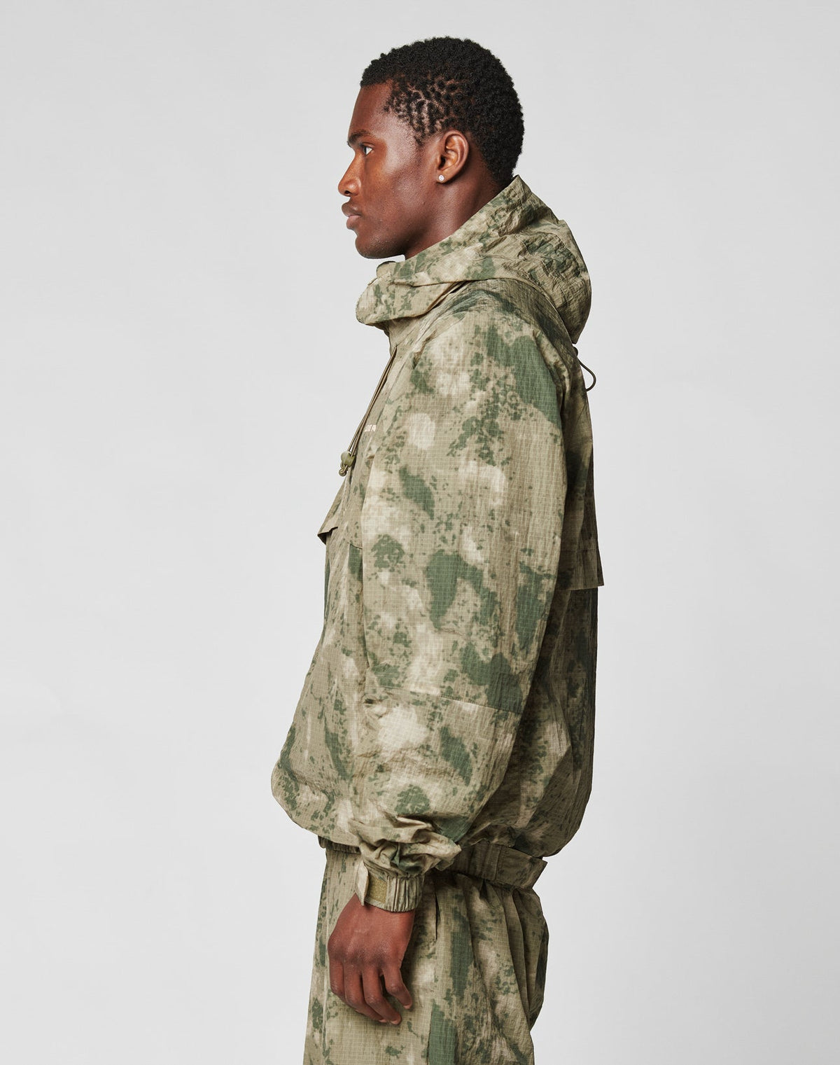A man in profile models the LIVE FAST DIE YOUNG Live Fast Nylon Jacket in oversize green and beige camo with a hood and matching pants, standing against a plain light gray background.