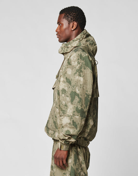 A man in profile models the LIVE FAST DIE YOUNG Live Fast Nylon Jacket in oversize green and beige camo with a hood and matching pants, standing against a plain light gray background.