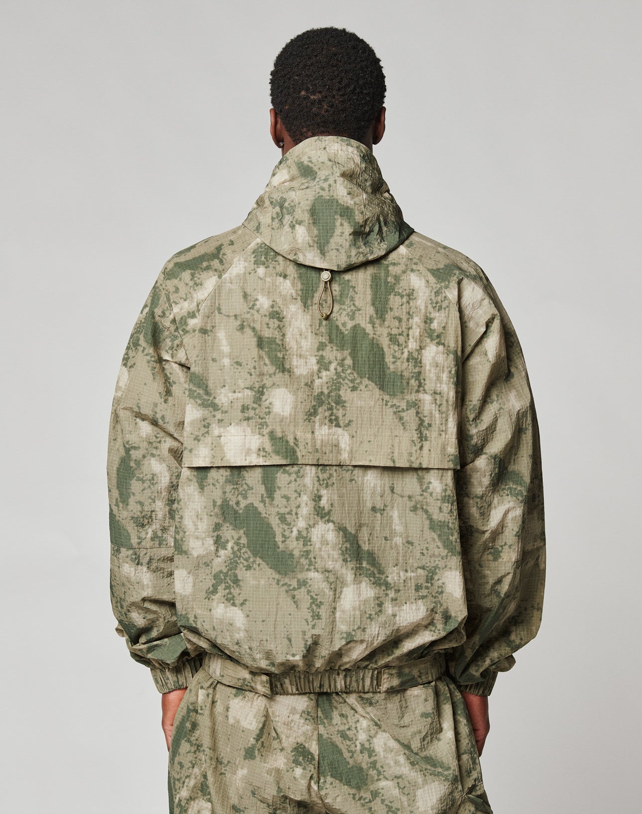A person stands facing away, wearing the LIVE FAST DIE YOUNG Live Fast Nylon Jacket in light green and beige camo with a hood and matching pants, hands at their sides against a plain light background.