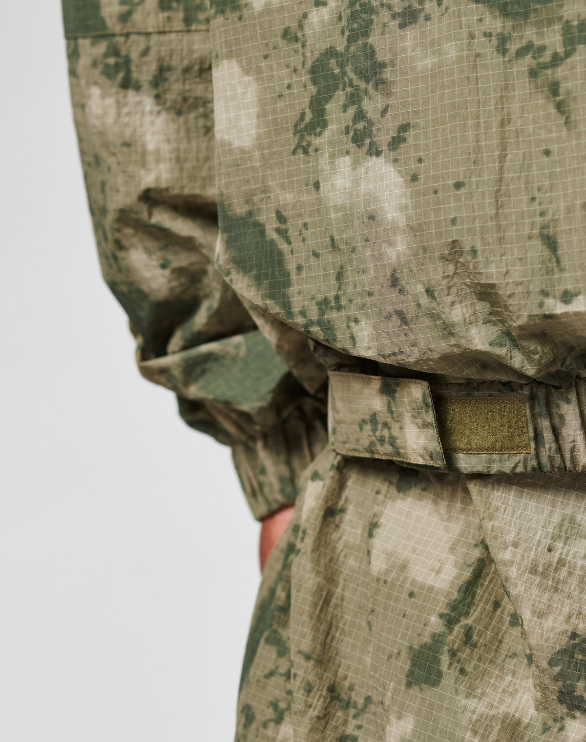 A close-up of a person wearing the Live Fast Nylon Jacket by LIVE FAST DIE YOUNG in green and beige digital camo, highlighting the sleeve, waistband, and zippered pockets. The background is plain and light.
