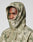 A person wears the Live Fast Nylon Jacket by LIVE FAST DIE YOUNG, an oversized green and beige camo hooded jacket with elastic drawstring, hood up and zip covering the mouth, revealing only their eyes, nose, and part of the forehead.
