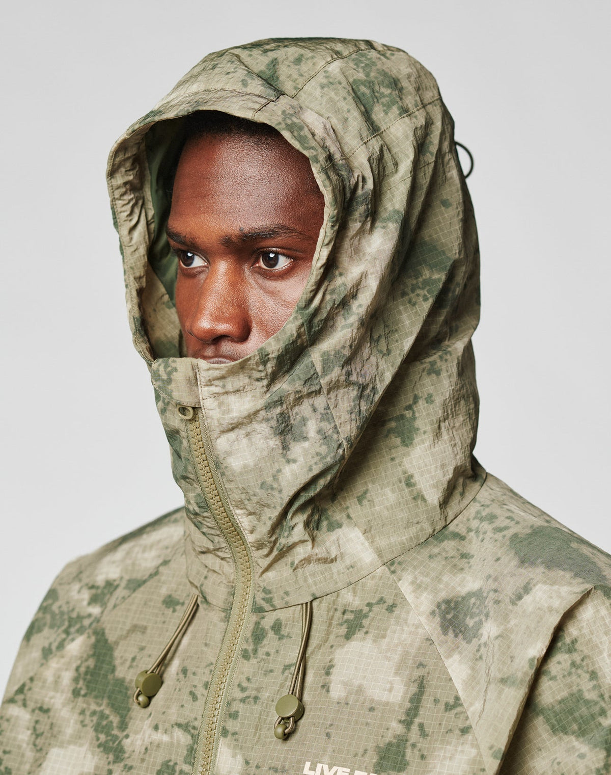 A person wears the Live Fast Nylon Jacket by LIVE FAST DIE YOUNG, an oversized green and beige camo hooded jacket with elastic drawstring, hood up and zip covering the mouth, revealing only their eyes, nose, and part of the forehead.