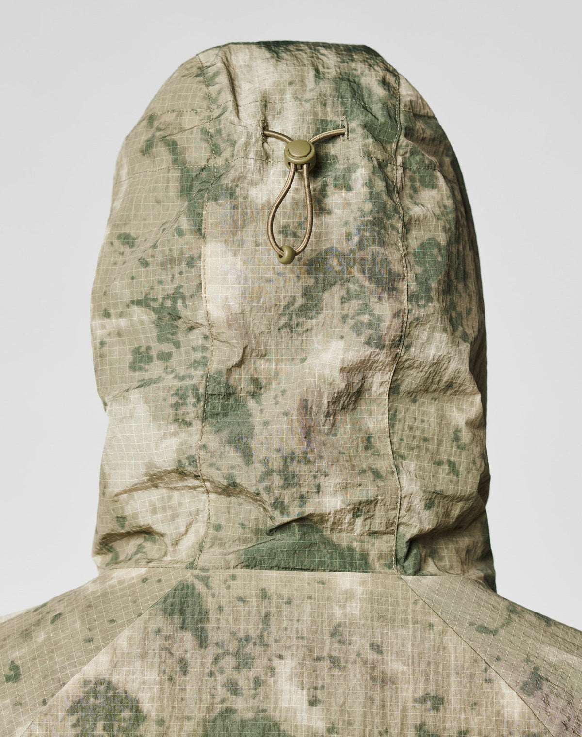 A close-up of the back of the LIVE FAST DIE YOUNG Live Fast Nylon Jacket in an oversize fit with green and beige camouflage, a hood with elastic drawstring and plastic toggle, and grid-textured fabric.