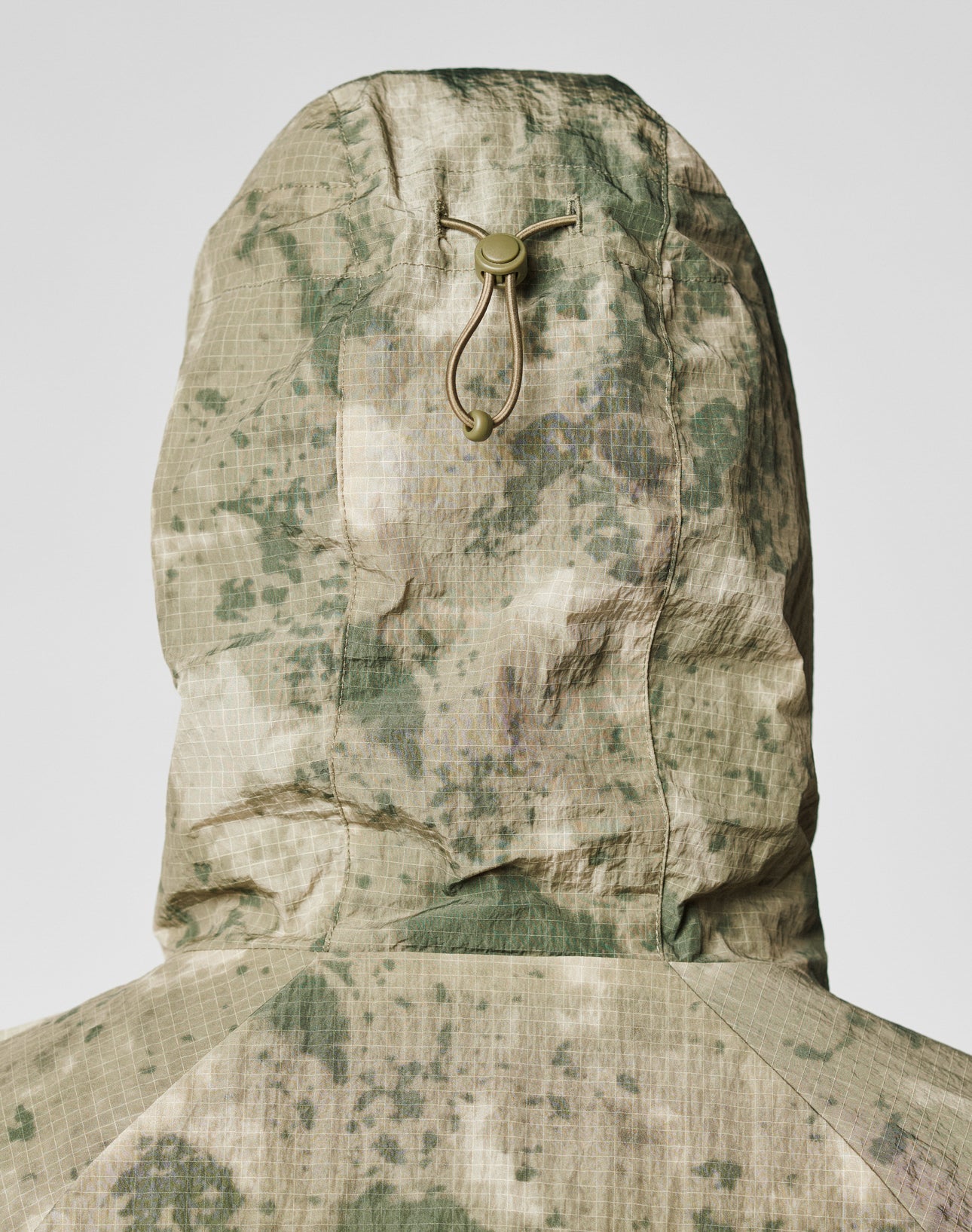 A close-up of the back of the LIVE FAST DIE YOUNG Live Fast Nylon Jacket in an oversize fit with green and beige camouflage, a hood with elastic drawstring and plastic toggle, and grid-textured fabric.