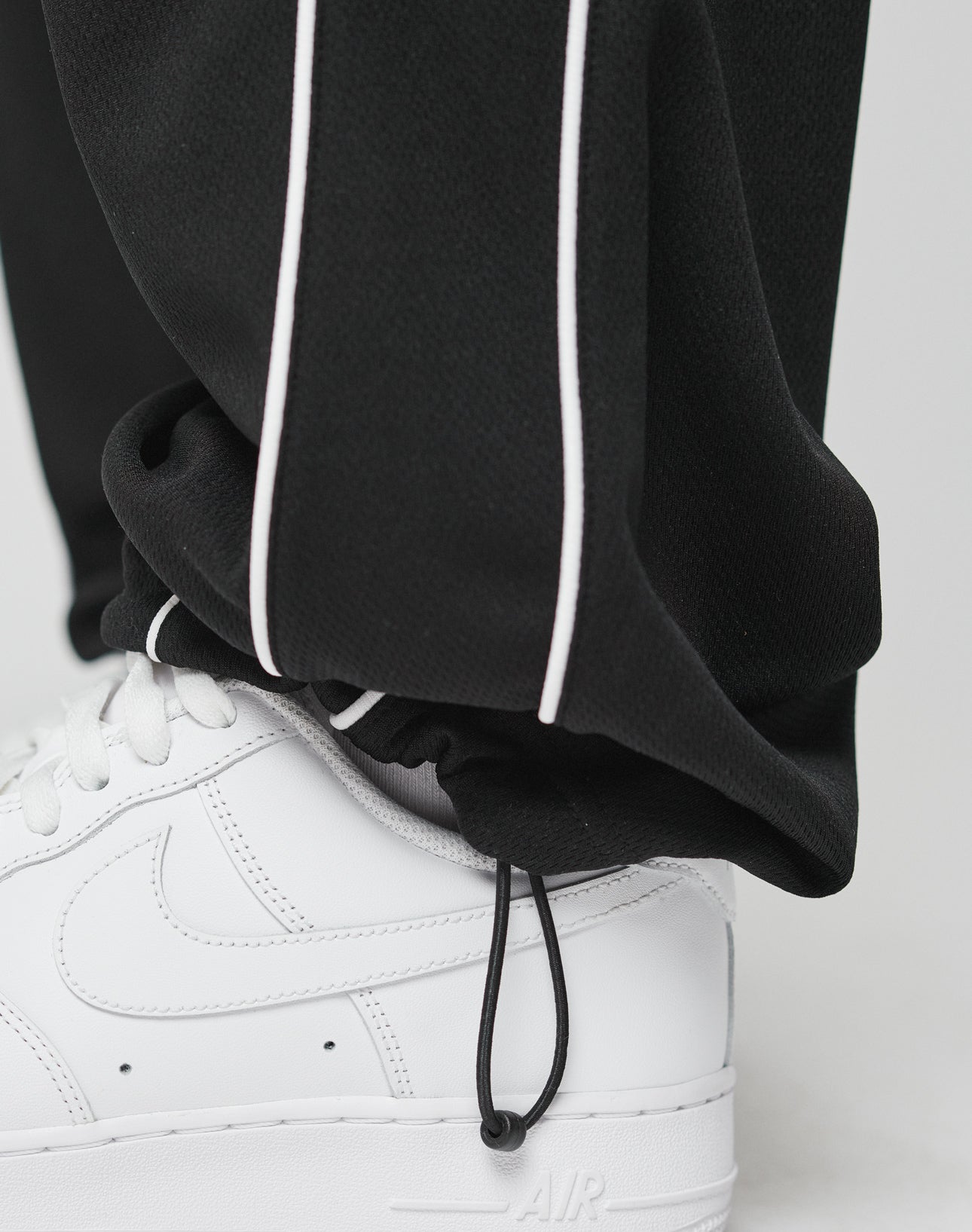A close-up of a person wearing LIVE FAST DIE YOUNG LFDY 55 Trackpants in wide fit black with white piping and an elastic waistband, paired with white Nike Air Force 1s. The polyester pants have a drawstring hem resting on the sneaker.