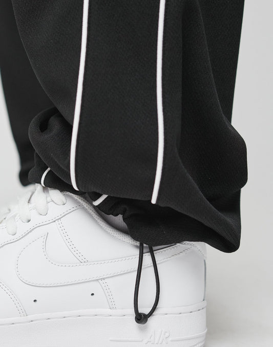 A close-up of a person wearing LIVE FAST DIE YOUNG LFDY 55 Trackpants in wide fit black with white piping and an elastic waistband, paired with white Nike Air Force 1s. The polyester pants have a drawstring hem resting on the sneaker.