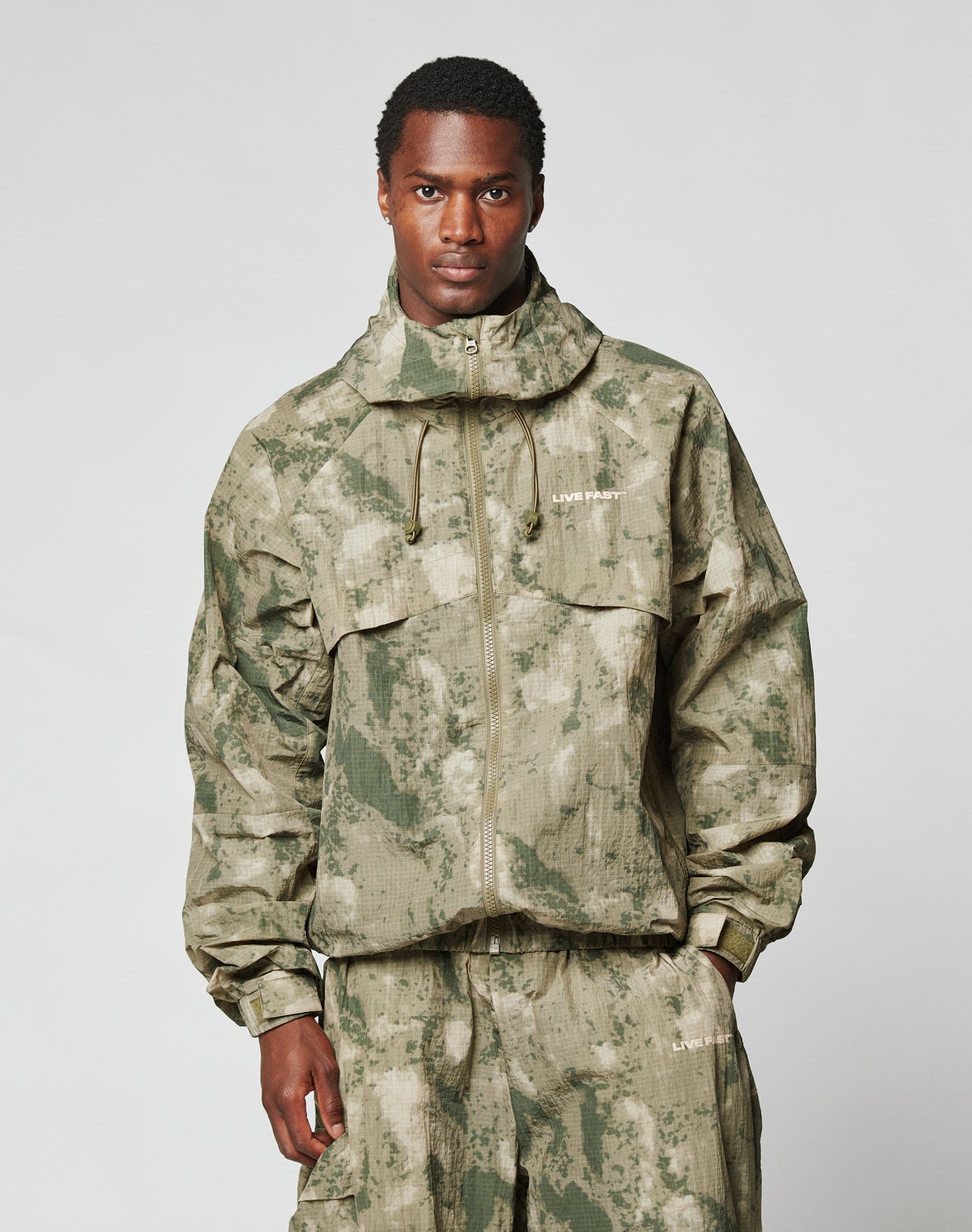 A man wears the LIVE FAST DIE YOUNG Live Fast Nylon Jacket in an oversize green and beige camouflage pattern with a hood and matching pants, standing facing the camera against a plain light background.