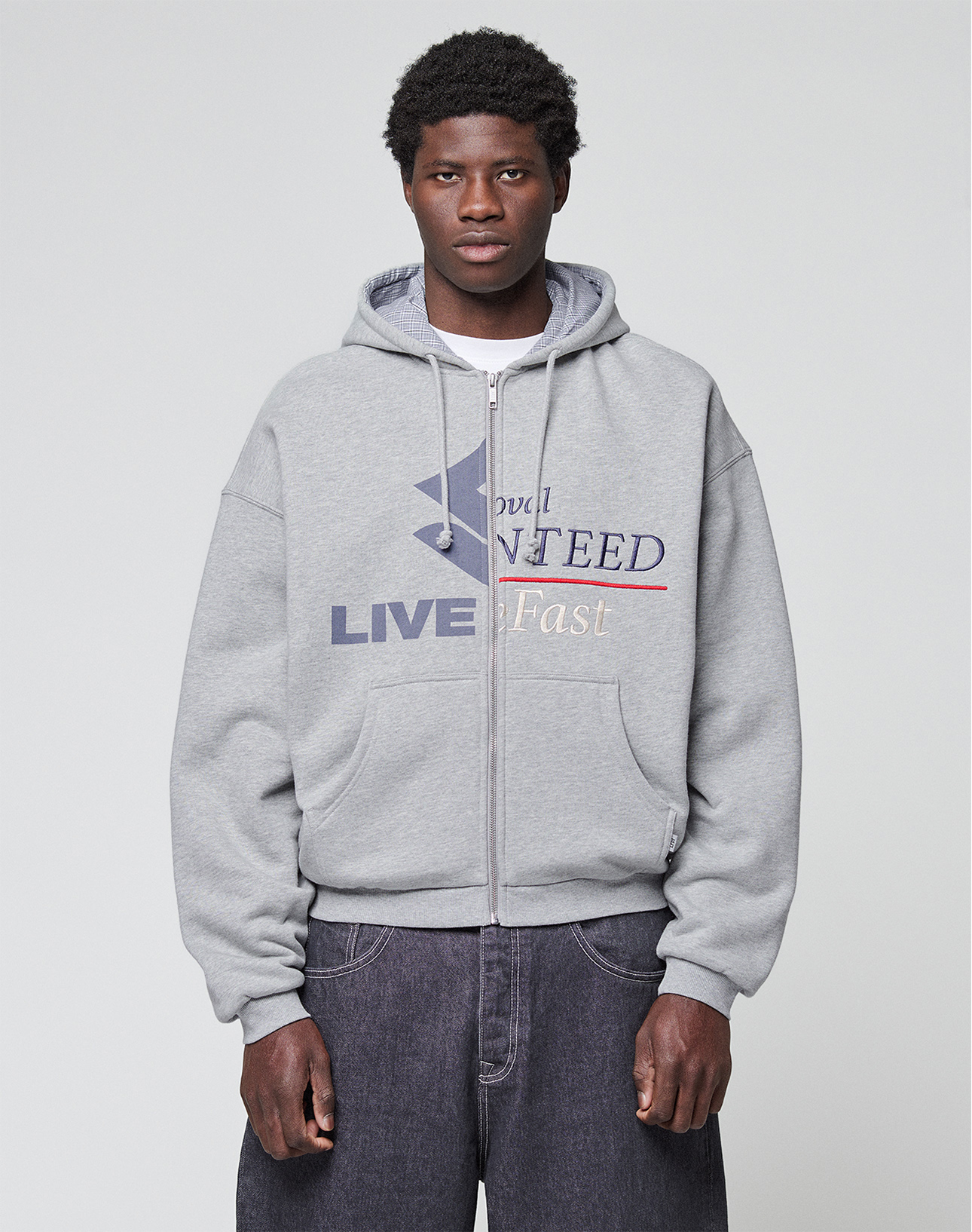 A person wearing a Flannel Lined Hooded Jacket by LIVE FAST DIE YOUNG, featuring a bold graphic and text, pairs it with loose dark gray jeans while standing against a plain light gray background.