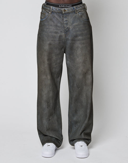 A person wearing LFDY Coated Baggy Denim by LIVE FAST DIE YOUNG—baggy fit, faded black with subtle wear, straight leg, white sneakers, and a black waistband featuring LIVE FAST in white. Crafted from comfortable Baumwolle.