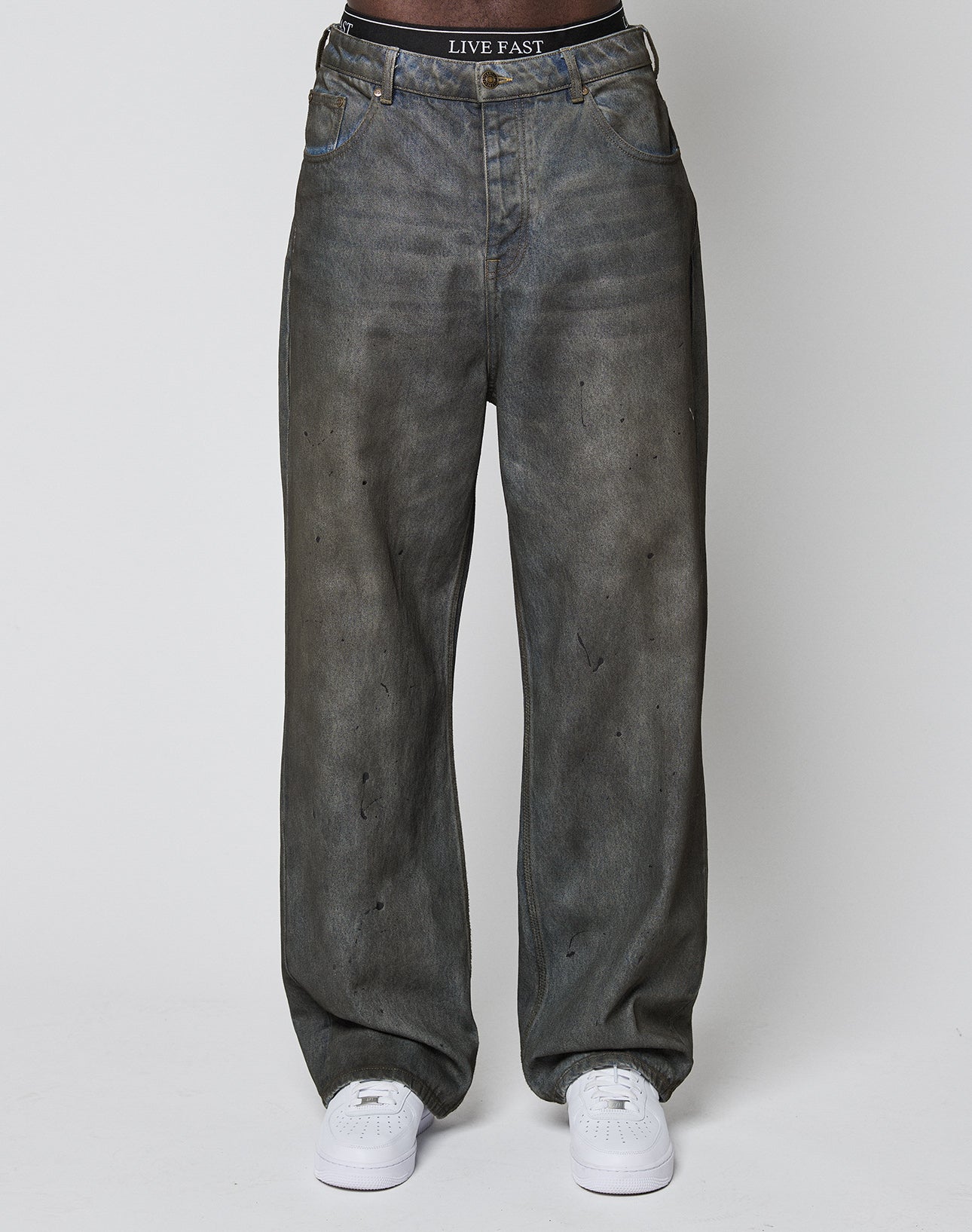 A person wearing LFDY Coated Baggy Denim by LIVE FAST DIE YOUNG—baggy fit, faded black with subtle wear, straight leg, white sneakers, and a black waistband featuring LIVE FAST in white. Crafted from comfortable Baumwolle.
