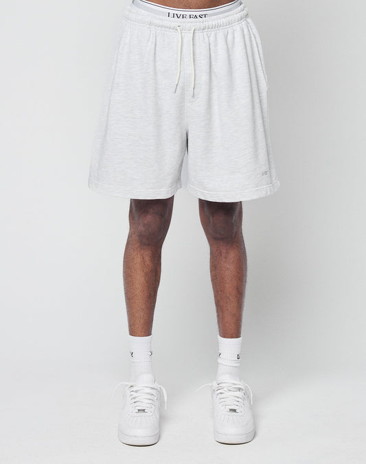 A person is shown from the waist down, wearing LIVE FAST DIE YOUNG Basic Sweat Shorts in light gray, paired with white socks and sneakers, standing against a plain light gray background.