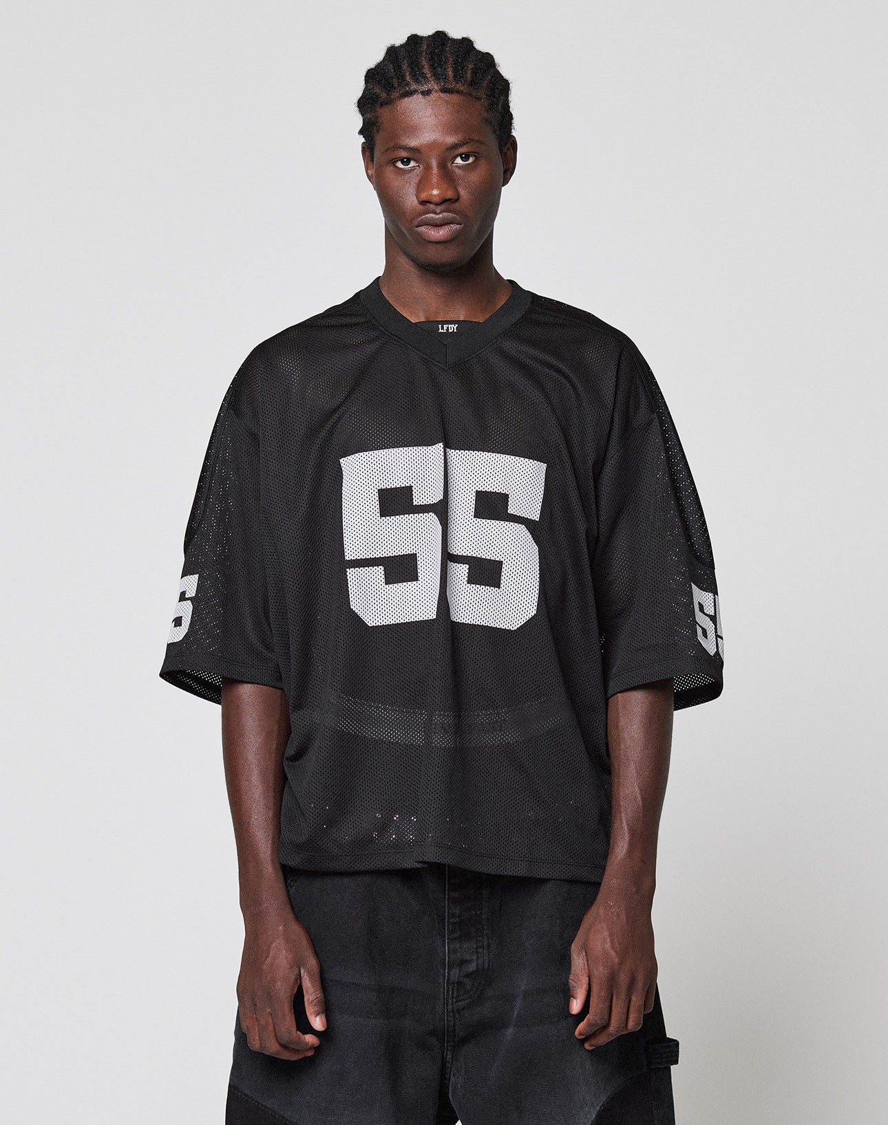 A person with braided hair wears an oversize black LFDY Mesh Jersey by LIVE FAST DIE YOUNG, featuring large white number 55 on the front and sleeves, standing against a plain light gray background. | model