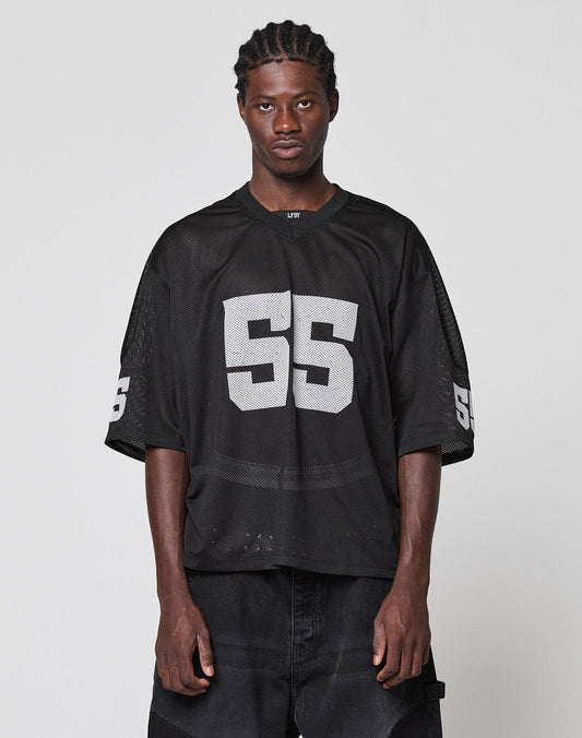 A person with braided hair wears an oversize black LFDY Mesh Jersey by LIVE FAST DIE YOUNG, featuring large white number 55 on the front and sleeves, standing against a plain light gray background. | model