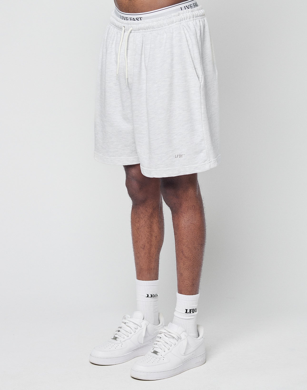 Someone wearing LIVE FAST DIE YOUNG Basic Sweat Shorts in light gray with an elastic waistband and subtle logo print, paired with white socks and sneakers, stands against a plain white background. Only their lower body is visible. | model