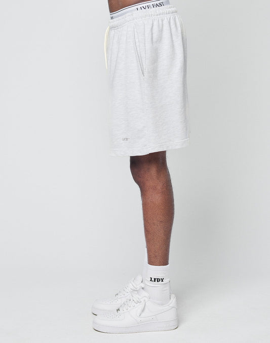 A person stands sideways against a plain background, showing only the lower half of their body in light grey LIVE FAST DIE YOUNG Basic Sweat Shorts with elastic waistband, white LFDY logo socks, and white sneakers.